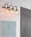 Lofty Chic Three Light Vanity Light In Dark Bronze With Clear Glass Shades And Antique Brass Accents Over Bathroom Mirror