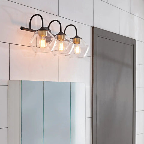 Lofty Chic Three Light Vanity Light In Dark Bronze With Clear Glass Shades And Antique Brass Accents Over Bathroom Mirror