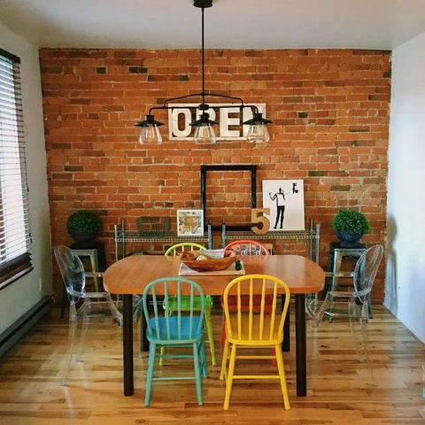 Dining area with Three Light Pendant In Oil Rubbed Bronze With Clear Glass Shades, vintage industrial vibe, above a wooden table and colorful chairs.