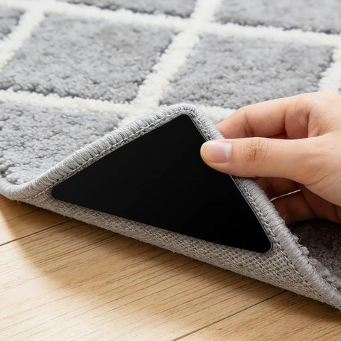 Hand applying Non Slip Rug Grippers 12 Pack Double Sided Adhesive Rug Tape Reusable Triangle Corner Pads under a grey rug on hardwood floor.