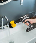 Hand holding Beast Of A 20V Max Cordless Drill Set With Lithium Battery with brush attachment near a running faucet.