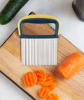Stainless steel crinkle cutter with blue-yellow ergonomic handle on cutting board, carrot slices nearby.