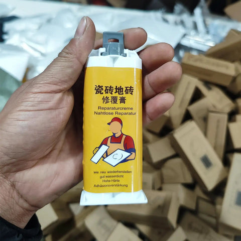 Hand holding Fast Dry Tile Repair Glue Kit For Marble And Granite That Delivers Seamless Crack Chip Hole Fix And Stone Restoration Diy Friendly Durable Non Toxic Formula Clear Cure Two Pack against a background of boxes.