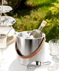 Double Wall Vacuum Insulated Ice Bucket with lid, scoop, ice tong, strainer, leather handle, and bottle on outdoor table.