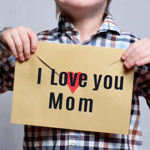 Child holding card with 'I Love You Mom' message, heart symbol, wearing plaid shirt.
