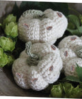 Crocheted white pumpkins with green stems in a decorative arrangement surrounded by green foliage