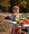 Child holding plate near Foldable Outdoor Picnic Camping Table With Adjustable Height Aluminum Legs And Roll Up Top, Silver Finish