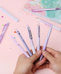 Hands holding Pastel Purple Gel Pen Set 12 Pack With One Matching Highlighter Black Ink 0.5mm Fine Point Smooth Gel Pens For Journaling School And Office Aesthetic Girly Stationery on desk.