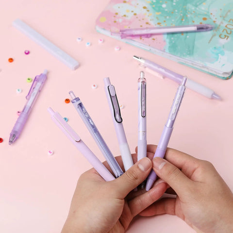 Hands holding Pastel Purple Gel Pen Set 12 Pack With One Matching Highlighter Black Ink 0.5mm Fine Point Smooth Gel Pens For Journaling School And Office Aesthetic Girly Stationery on desk.