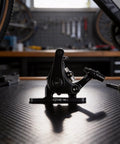 Kickass Mechanical Cable Disc Brake Set For Mountain And Road Bike Featuring Solid Aluminum Alloy Calipers and 160mm 140mm Adapters, side view of a black brake caliper on a workbench.