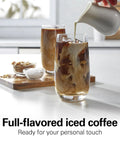 Pouring creamer into a glass of iced coffee on a kitchen counter with a wooden tray of ingredients nearby.