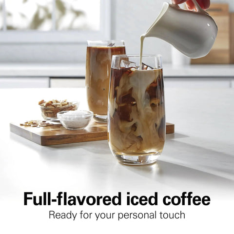 Pouring creamer into a glass of iced coffee on a kitchen counter with a wooden tray of ingredients nearby.