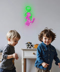 Two children playing under a green and pink neon alien sign on a bedroom wall.