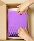 Hands placing Purple Self Seal Bubble Mailers 18x23cm, 10 Pack, Thickened Aluminum Foil Padded Envelopes into a box.