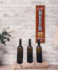 Rustic Cold Beer Served Here Metal Wall Sign Retro Vertical Tin Decor mounted on brick wall near brown bottles and lamp.
