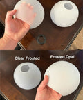 Person holding frosted opal and clear frosted glass globe, part of Three Pack Frosted Opal White Glass Globes Replacement For Light Fixtures 5.9 Inch Diameter 5 Inch Tall Soft White Round Lampshade Cover For Pendant Lights Wall Sconces And Chandeliers One And Five Eighth Inch Fitter E26 Bulb Compatible.