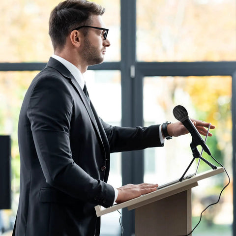 Man standing at podium with 800 Pcs Disposable Microphone Covers Non Woven with Elastic Band Individually Wrapped Mic Covers for KTV Karaoke Studio Recording Stage Black for Stage Use microphone cover on mic.