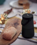Revolve Plus II Portable Bluetooth Speaker with 360 Degree Sound and Built in Microphone beside bread on a wooden table.