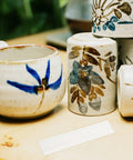 White ceramic pottery cups and jars with blue floral glaze on a wooden table; white ribbed strip in foreground