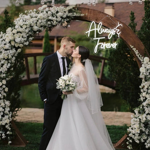 Wedding couple kissing beneath floral arch with 'Always & Forever' neon sign