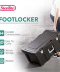 Flex Your Storage Game With Heavy Duty Plastic Footlocker Rolling Bin With Wheels And Handle, Stackable Tote Featuring Secure Latching Lid For Basements Dorms And Shared Spaces, Smooth Operator Hauler Chest In Black, durable black bin being carried.