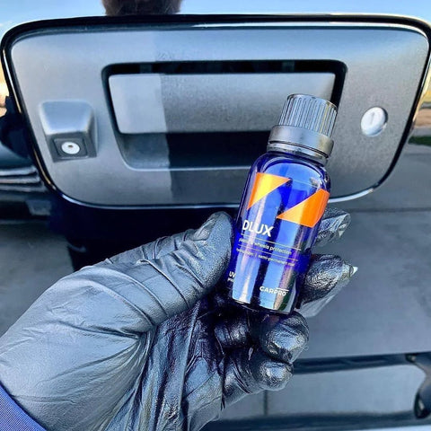 CARPRO DLUX wheel and trim coating bottle (blue with orange label) held by a gloved hand in front of a car tailgate.