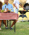 Outdoor picnic scene featuring a table with red checkered tablecloth, wine, and food beside a barbecue grill.