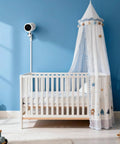White crib with canopy in blue nursery room, camera on wall for baby monitoring, natural light casting shadows on wall.