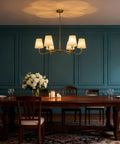 Smooth Operator 6 Light Brass Gold Chandelier With White Fabric Shades hung over a wooden dining table with flower centerpiece.