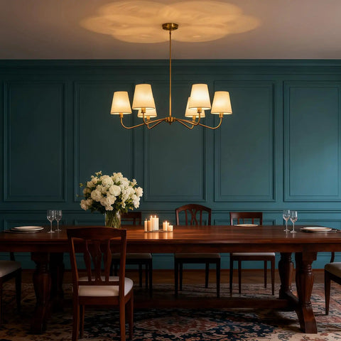 Smooth Operator 6 Light Brass Gold Chandelier With White Fabric Shades hung over a wooden dining table with flower centerpiece.