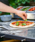 Ooni 16-inch metal pizza peel with long handle sliding a basil-topped pepperoni pizza on an outdoor grill.