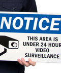 24 Hour Video Surveillance Security Camera Sign 10 By 14 Inch Heavy Duty Plastic Outdoor Notice Plaque held by a person.