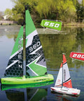 Two 1:22 Scale Wind Powered RC Racing Sailboats on a pond, green and white hull with mast and sails attached, trees in background