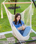Woman relaxing in Caribbean Style Hammock Chair With Spreader Bar, XL Rope Hanging Chair For Indoor Outdoor Use, 300 Lb Weight Capacity, Natural White, Cozy Swing For Garden, Patio, Or Backyard, Easy Setup, reading a book in a garden setting.