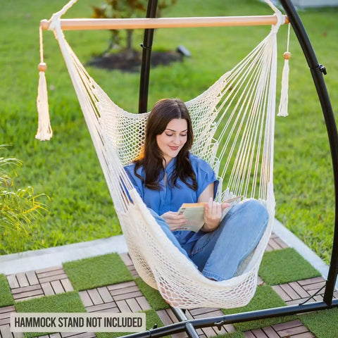 Woman relaxing in Caribbean Style Hammock Chair With Spreader Bar, XL Rope Hanging Chair For Indoor Outdoor Use, 300 Lb Weight Capacity, Natural White, Cozy Swing For Garden, Patio, Or Backyard, Easy Setup, reading a book in a garden setting.