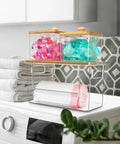 Kicking Clutter Away Trash Bag Dispenser 2 Pack, transparent acrylic containers with bamboo lids holding pods beside towels.
