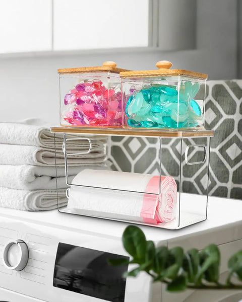 Kicking Clutter Away Trash Bag Dispenser 2 Pack, transparent acrylic containers with bamboo lids holding pods beside towels.