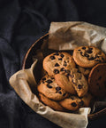 Bowl of chocolate chip cookies on parchment-lined bowl on dark cloth.