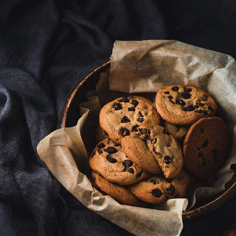 Bowl of chocolate chip cookies on parchment-lined bowl on dark cloth.