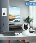 12V 4A 48W AC DC Power Supply Adapter powering indoor camera via 5.5x2.5mm jack, plugged into wall outlet in living room.
