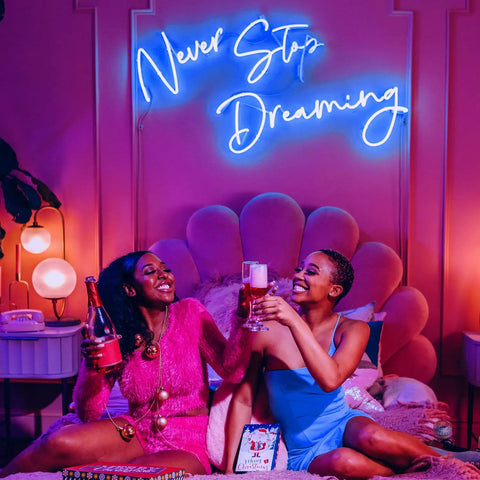 Two people sitting under a neon sign reading 'Never Stop Dreaming' in a stylish bedroom setting with colorful decor.