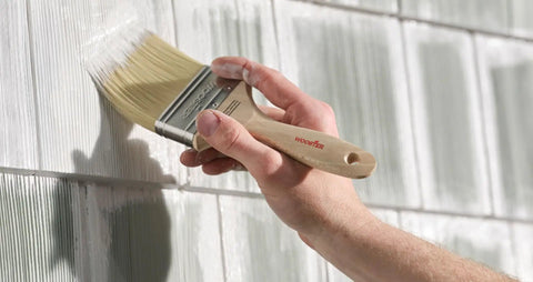 Person using a beige paintbrush on a textured surface, not a 2 Inch Shortcut Angle Sash Paintbrush Pack Of 3 With Purple Soft Grip Handle