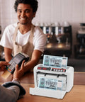 Portable Digital Money Counter With UV And MG Detection, 800 Bills Per Minute in use at a cafe counter, person holding card reader.