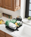 Compact Dish Drying Rack For Kitchen Counter Black Space Saving Drainer holds plates and bowls on a countertop next to a sink.