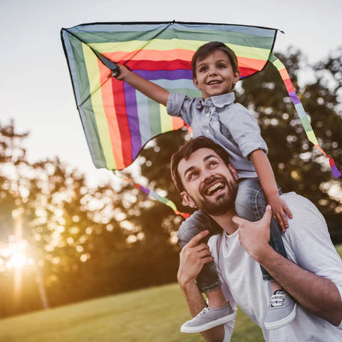 Father carrying son on shoulders flying a colorful kite at sunset in the park.