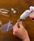 AdTech 4-inch mini hot glue gun applying clear glue to a glass ornament, with glue sticks nearby.