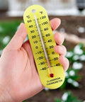 Hand holding a yellow classroom thermometer with dual Fahrenheit and Celsius scales, PVC material, against a blurred background.