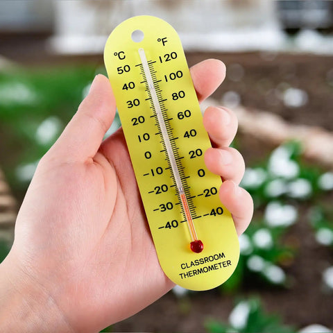 Hand holding a yellow classroom thermometer with dual Fahrenheit and Celsius scales, PVC material, against a blurred background.