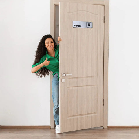 Woman smiling next to aluminum restroom sign for women on door; Two Pack Aluminum Restroom Signs For Men And Women Self Adhesive Metal Bathroom Door Wall Signage For Offices Restaurants Public Places 9 By 3 Inches Silvery Finish Easy Mount