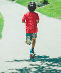 Child riding a kick scooter on a sunny park path; red shirt and blue shorts.